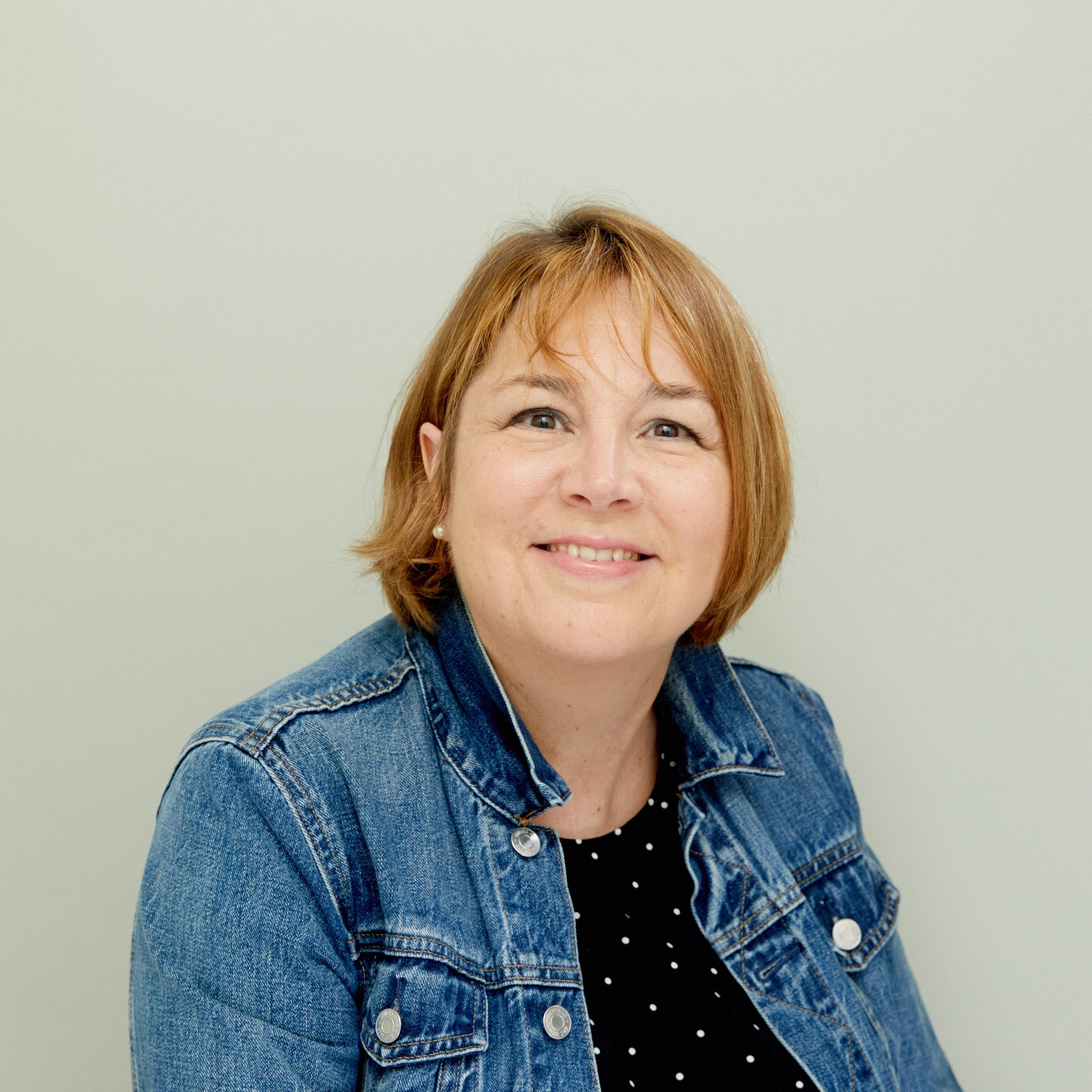 Portrait of a woman with short, light brown hair wearing a denim jacket and a polka dot top, sitting in front of a light green background.